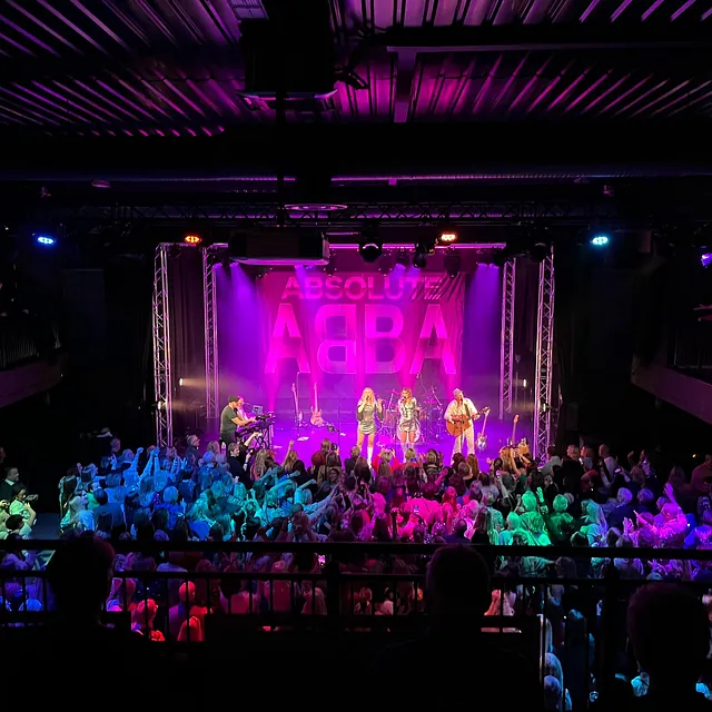 Band opptrer på scene med “Absolute ABBA”-banner bak, foran et entusiastisk publikum.