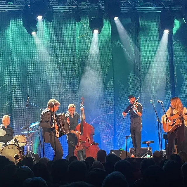 Band spiller live på scene med blå belysning foran et engasjert publikum i konsertsal.