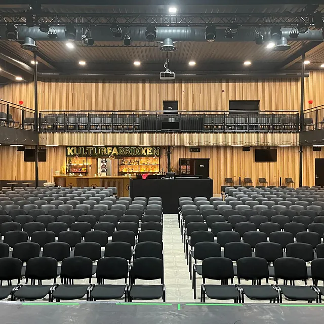 Stor sal med svarte stoler vendt mot scene, baren og galleriet til Kulturfabrikken i bakgrunnen.