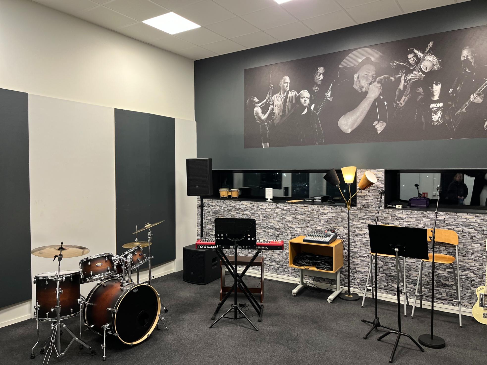 Musikkøvingsrom med trommesett, tangentinstrumenter og veggdekor med konsertbilder.