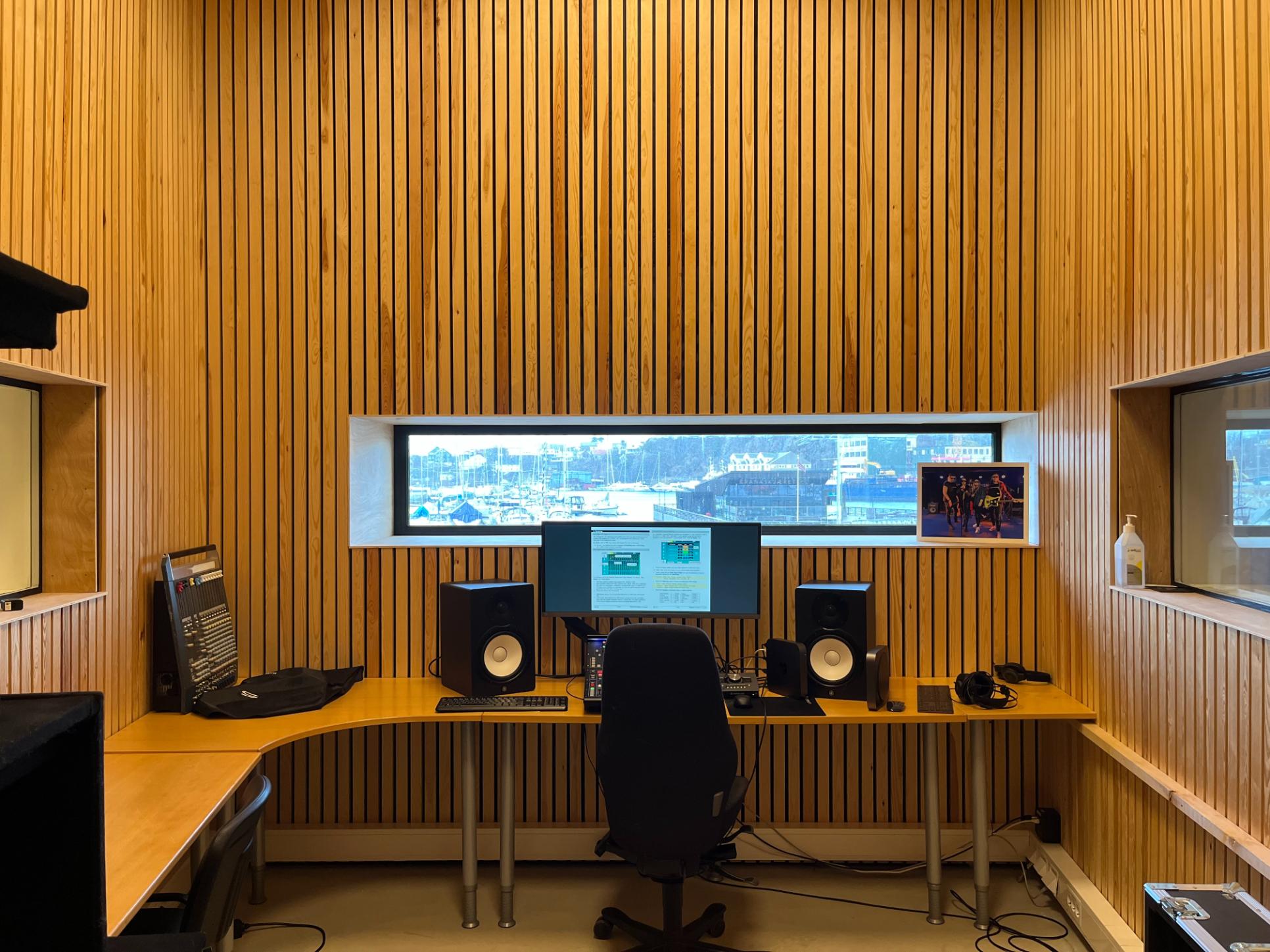 Lydstudio med trepanel, skjermer og studioutstyr, vindu med utsikt over havneområdet.